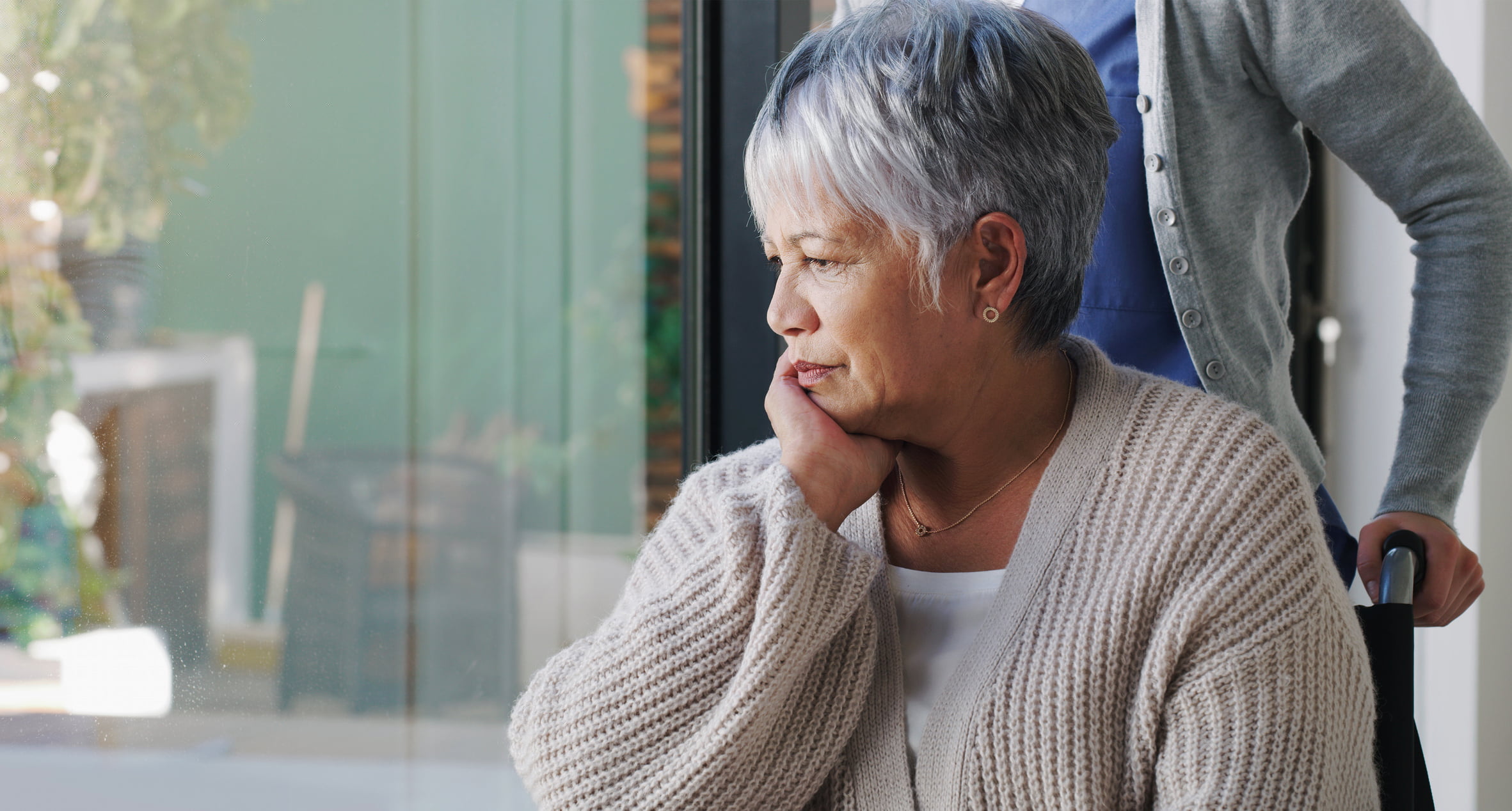 A pensive woman sits in her wheelchair as her caregiver holds the handles to push her